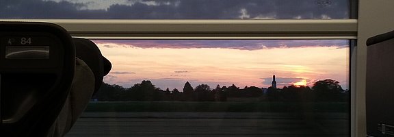 © Ralf Jankowski / fundus-medien.de Durch das Zugfenster eines schnell fahrenden Zuges sind man in der Ferne die Silhouette eines Kirchturms.